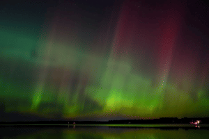 A photo of the northern lights, mostly greens and reds, reflecting over a lake
