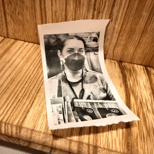 A black and white photograph of Hannah A. Patellis wearing a mask printed on receipt paper