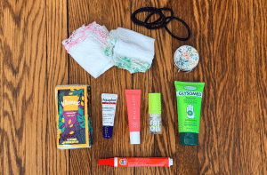 Nine small toiletry items are placed on a wooden floor. Hand cream, lip products, a Tide stick, pill case, Kleenex, 2 handkerchiefs, and hair ties
